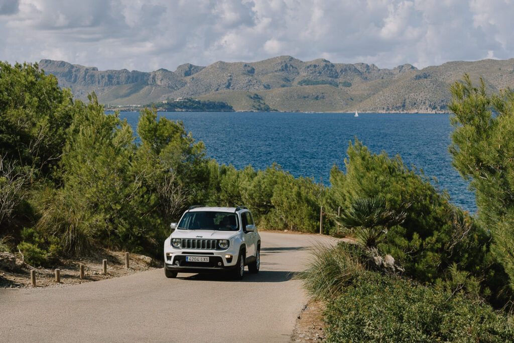 White jeep with hire car excess insurance driving up a hill with green trees either side and a blue sea behind.