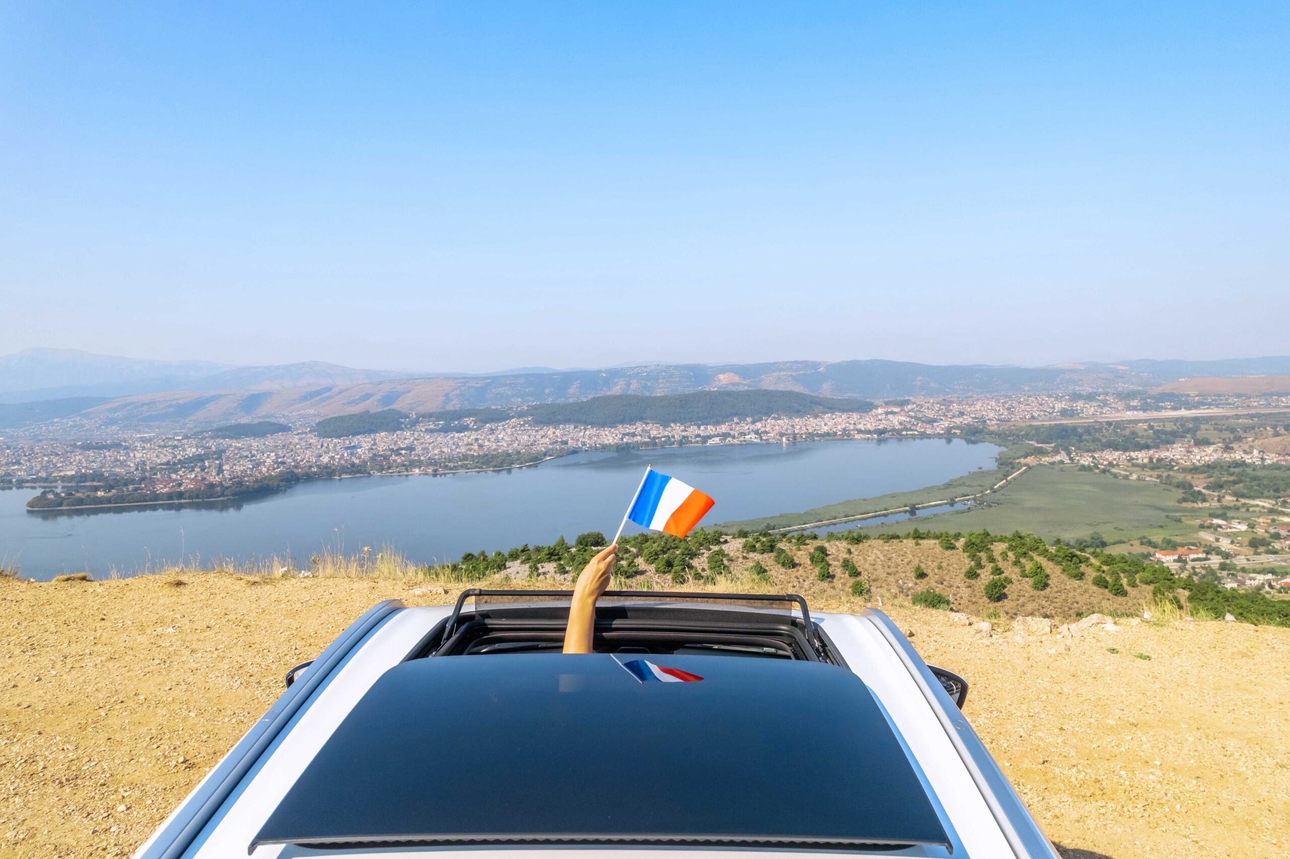 A hand waves a French flag from a car's sunroof with a serene lake and hills in the background to demonstrate why people hiring a car abroad should get car rental excess insurance.