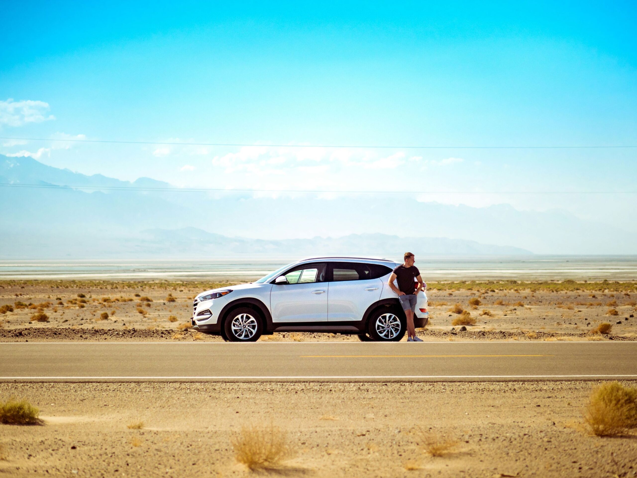 A person leans against a white SUV parked on a deserted road in a vast desert landscape.