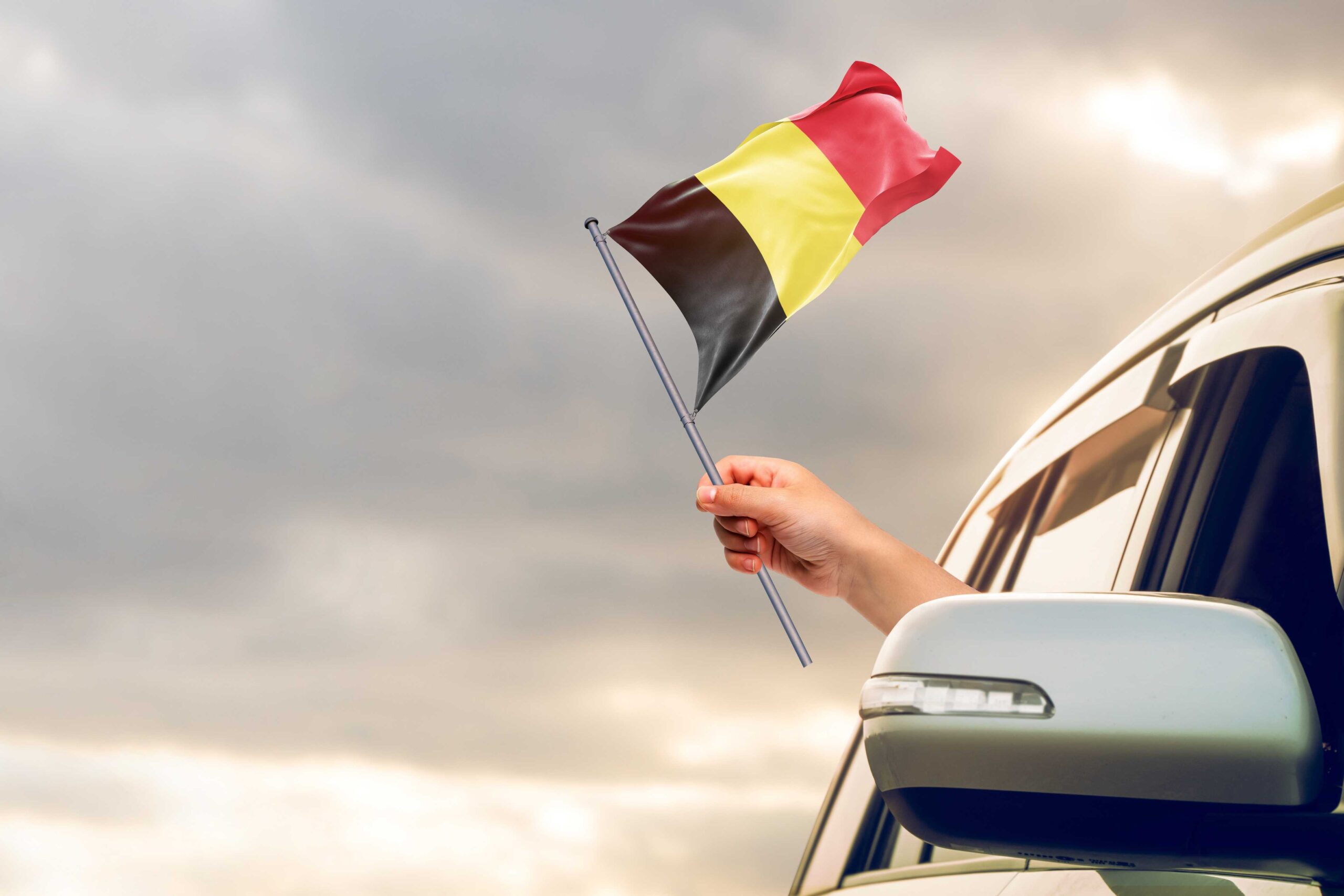 Person waving a Belgian flag out a car window with the cloudy sky behind to reinforce the importance for people renting cars to get car rental excess insurance.