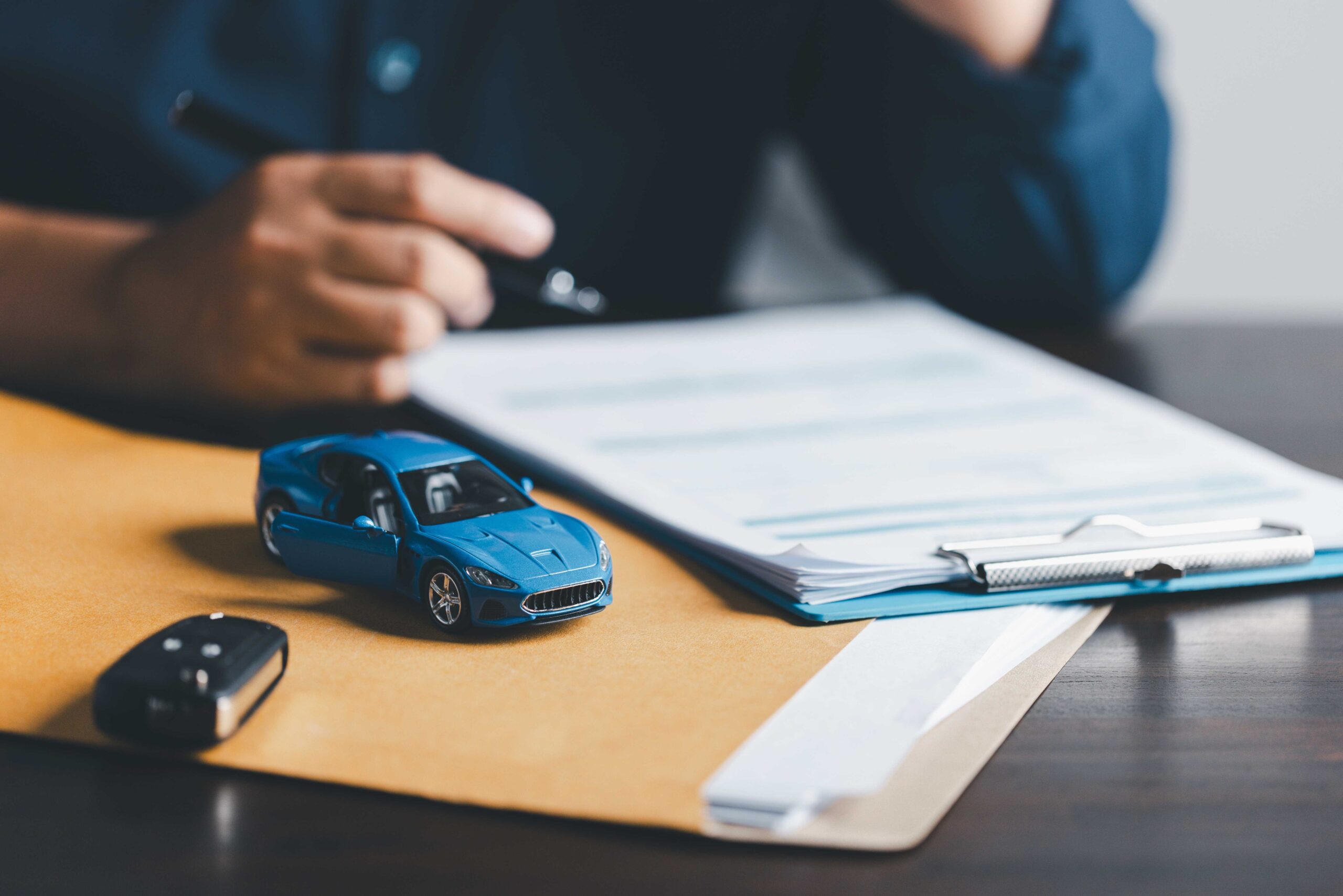 Person signing a car rental excess insurance agreement document, after purchasing car attached to clipboard with blue toy car in forefront.