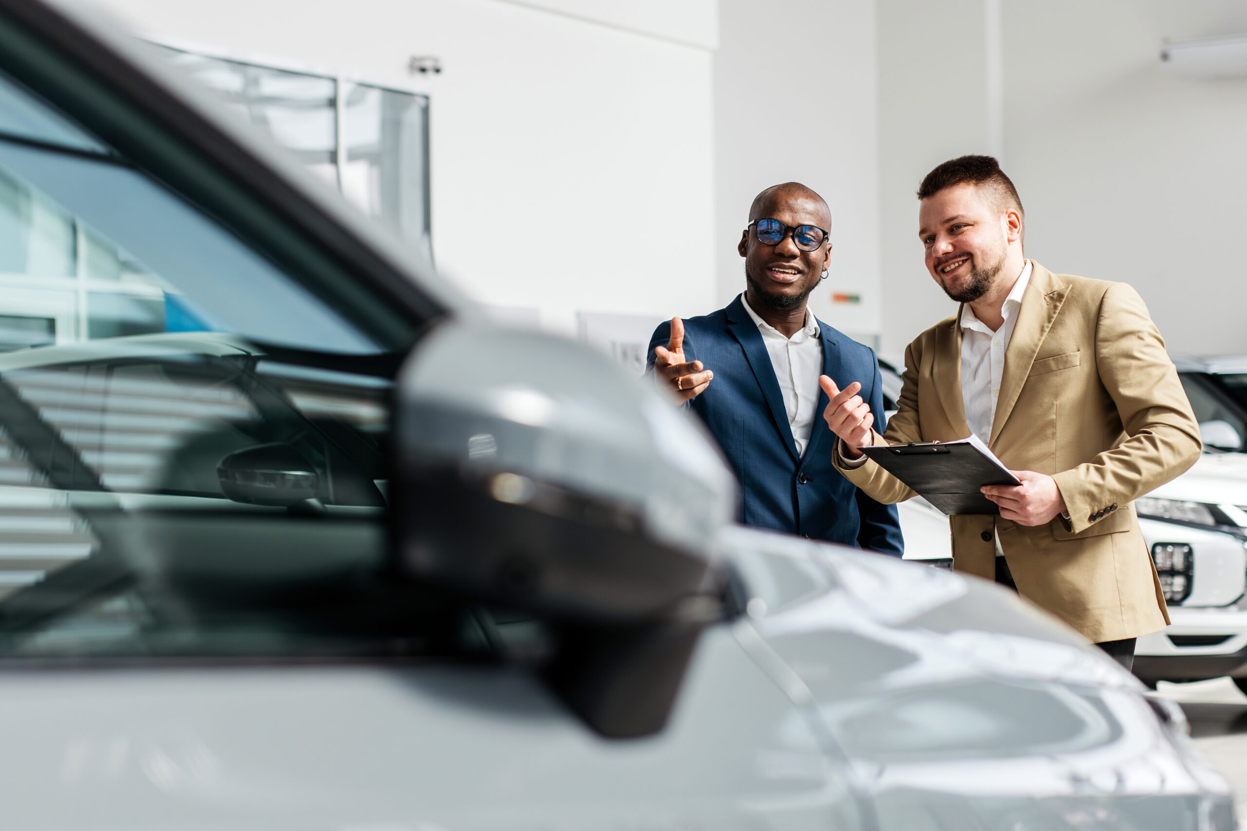 Grey car in the foreground with a salesperson and a client discussing it in the background.